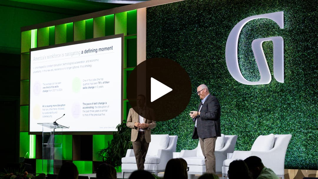 Two men present on a stage with green backlighting, next to a screen displaying slides. An audience watches from the foreground.