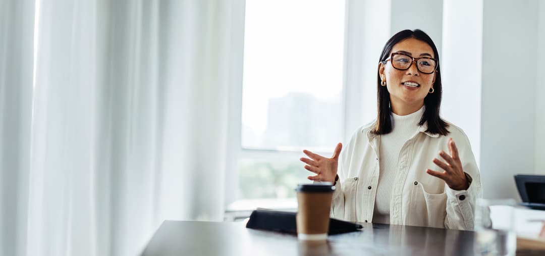 A woman with glasses talks animatedly at a desk with a coffee cup nearby, against a backdrop of large windows and white curtains.